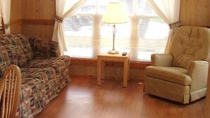 A cozy room with a wooden ceiling, a large window with curtains, a sofa, a chair, and a small table with a lamp.
