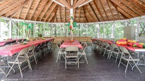 A covered outdoor dining area with multiple tables and chairs, a central decorative column, and a view of trees through the open sides.