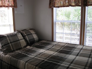 A bed with a plaid bedspread and two pillows, a window with blinds, and a wall with a bulletin board.