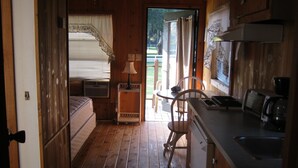 A compact kitchen with a dining area, a small table, and a chair. There is a bed with a window and curtains, and an air conditioning unit is visible.