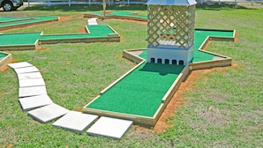 A miniature golf course with a small pavilion, green artificial turf, and a white fence.
