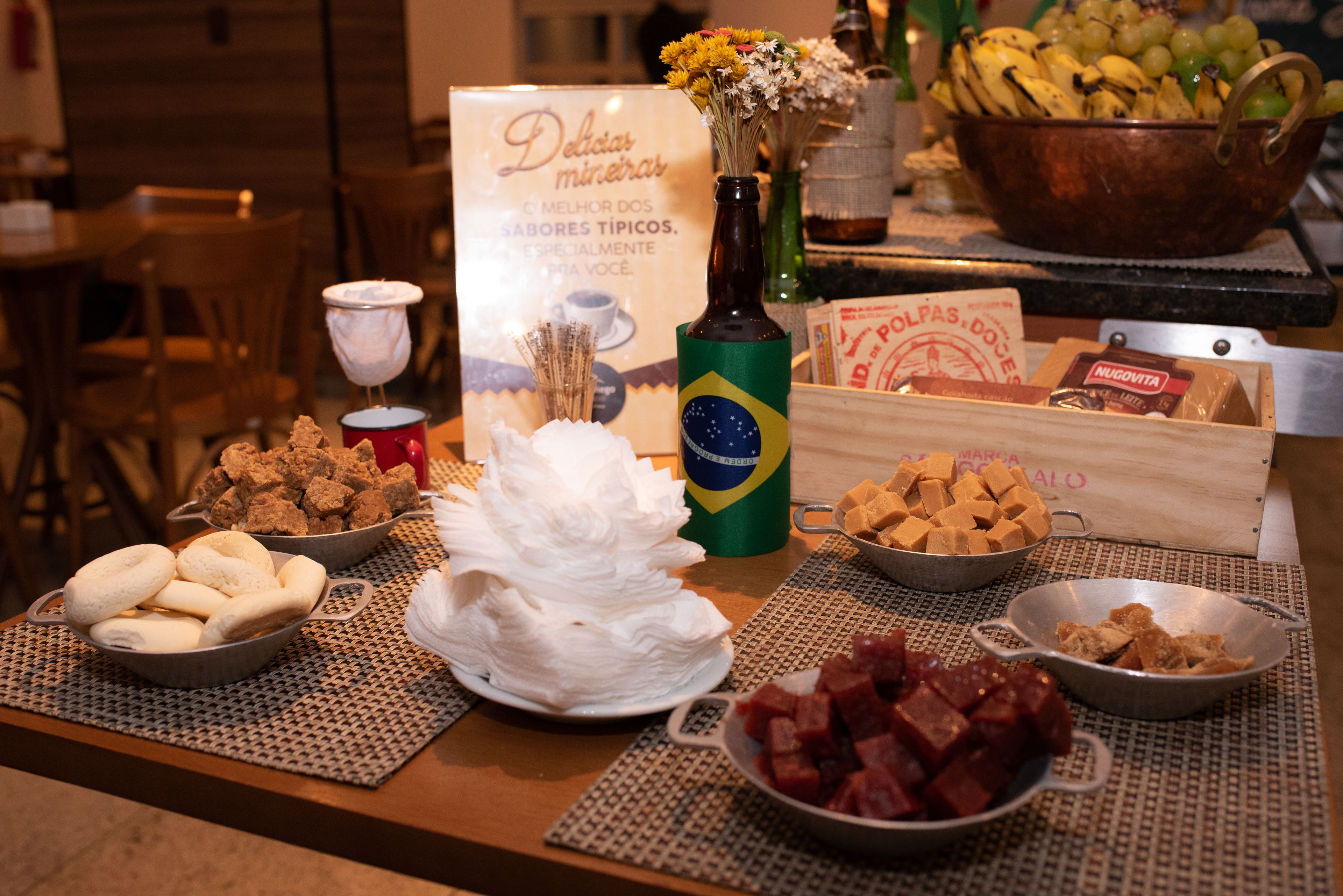Uma mesa de buffet com diversos alimentos, incluindo pão, doces e uma garrafa de cerveja brasileira.