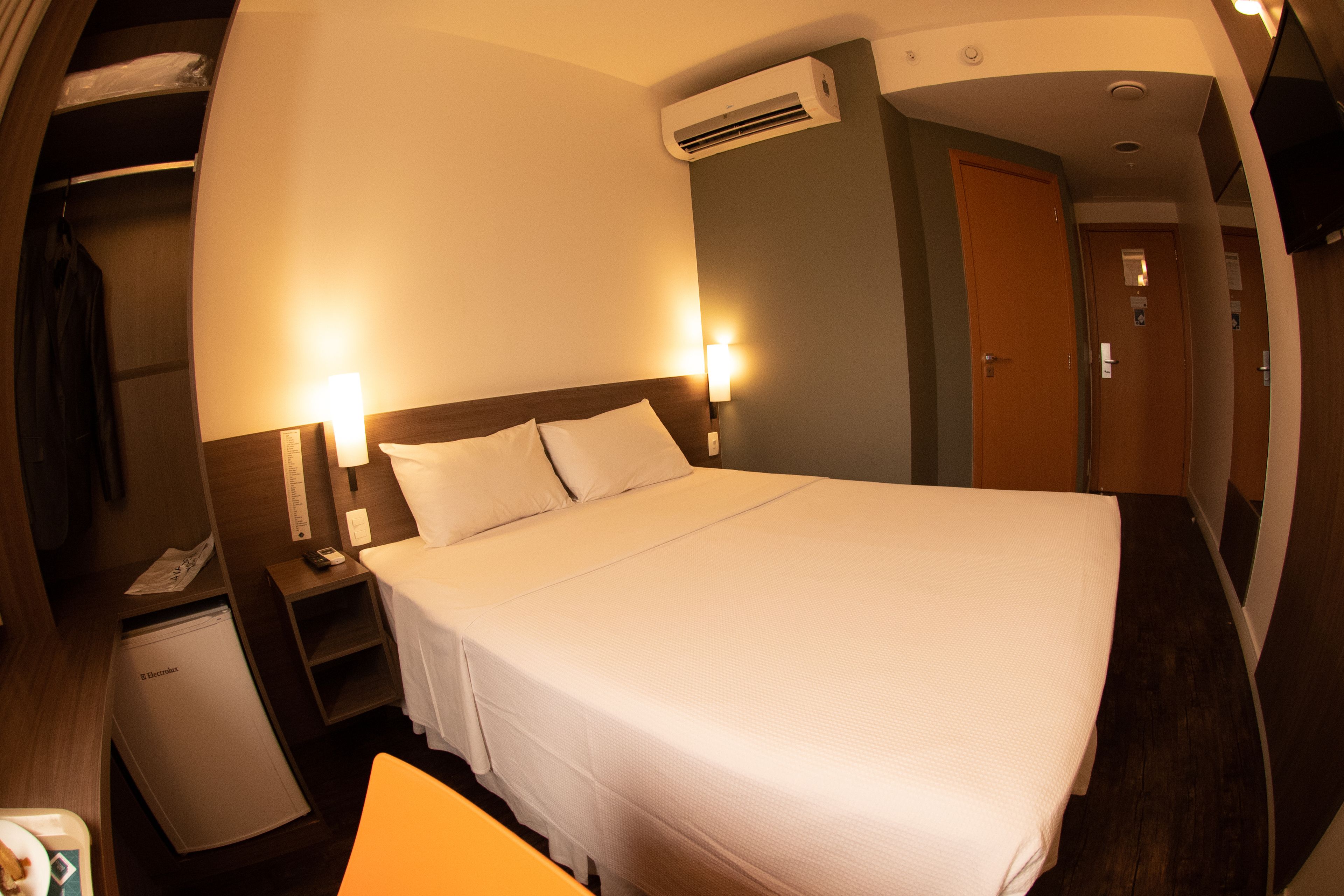 A hotel room with a bed, bedside lamps, a small desk with a phone, a mini-fridge, and an air conditioning unit.