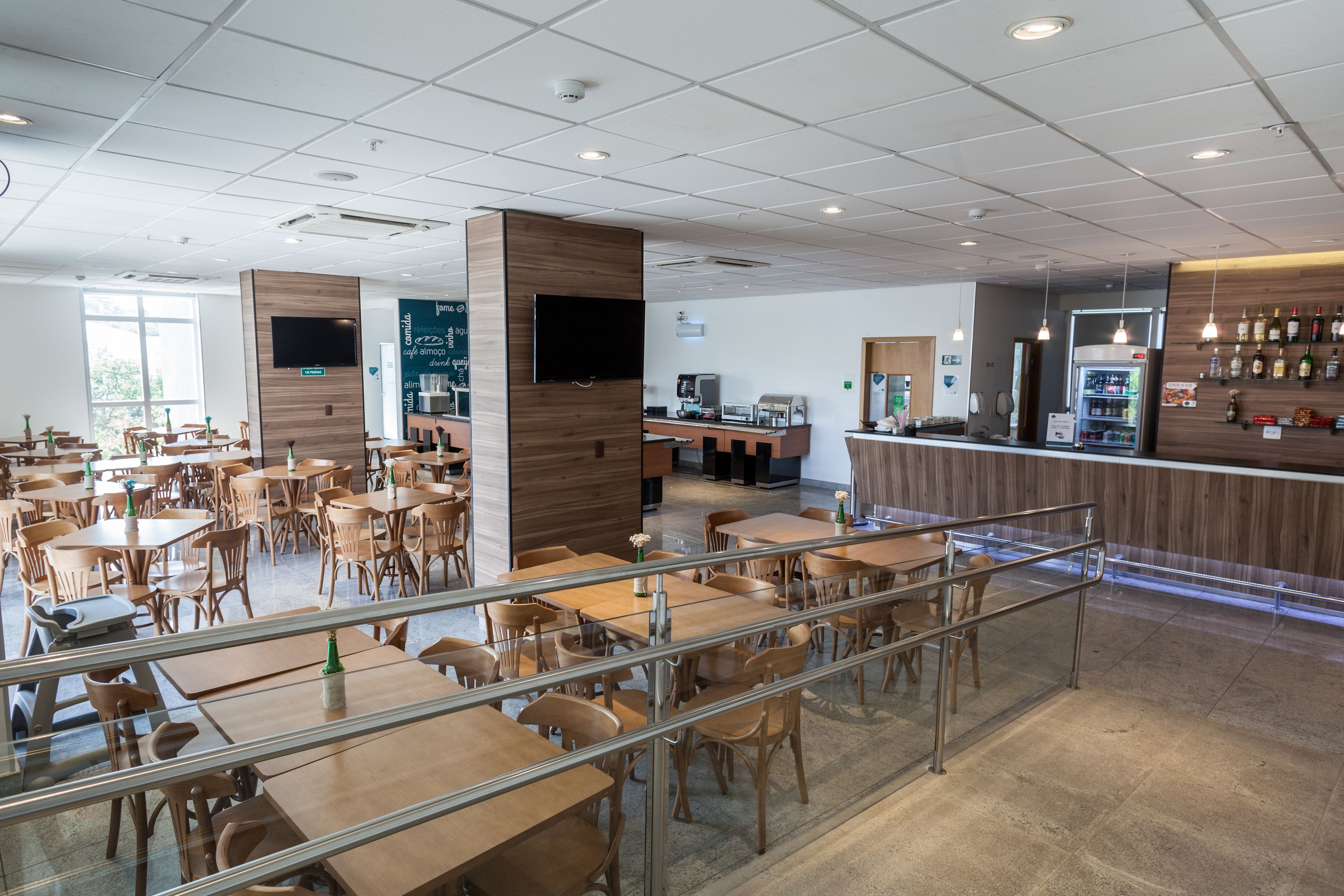 A spacious cafe with multiple tables and chairs, a counter with a beverage dispenser, and a wall-mounted TV.