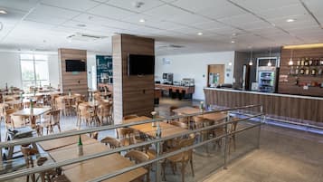 A spacious cafe with multiple tables and chairs, a counter with a beverage dispenser, and a wall-mounted TV.