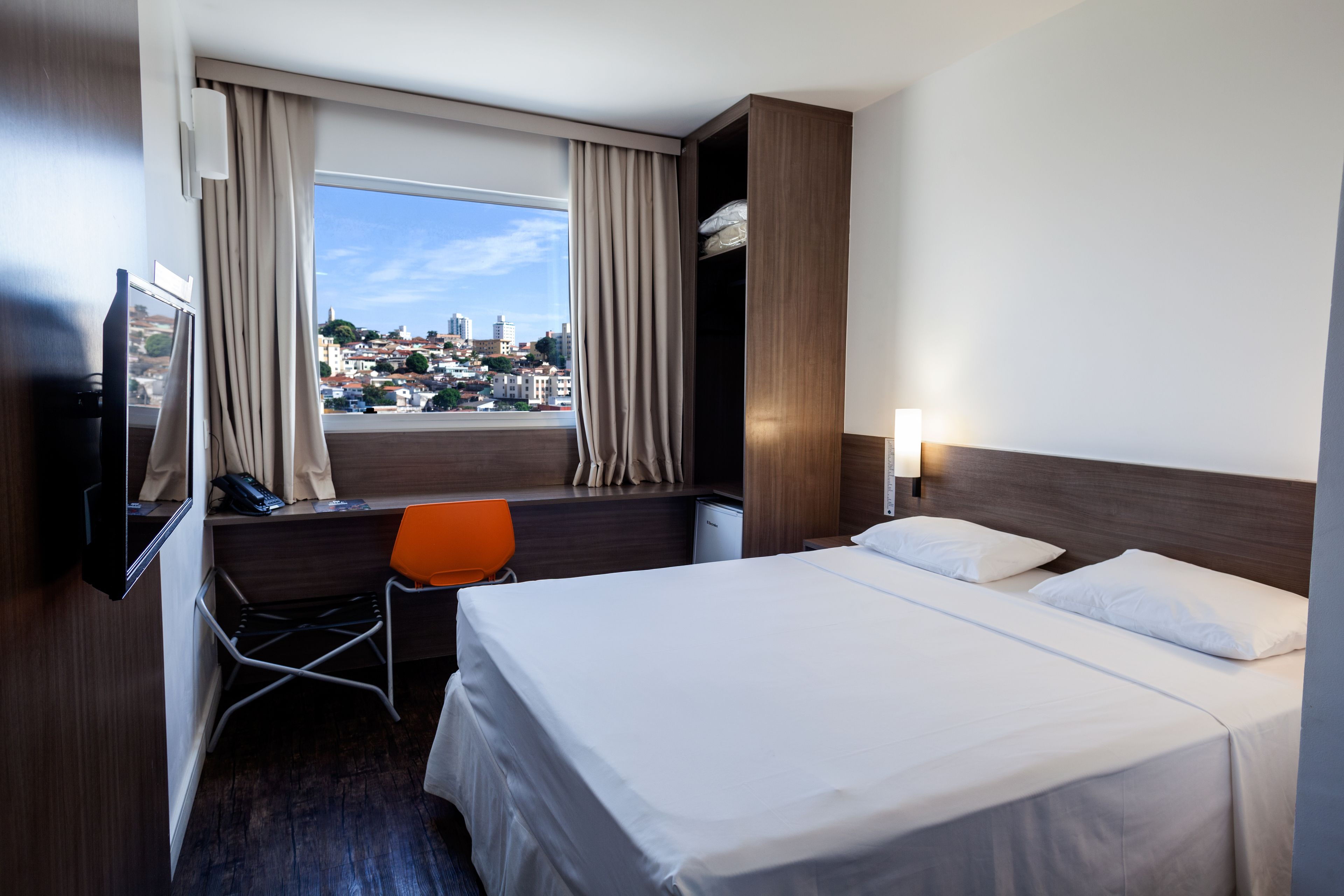 Quarto de hotel com cama, secretária com cadeira, televisão e janela com cortinas.