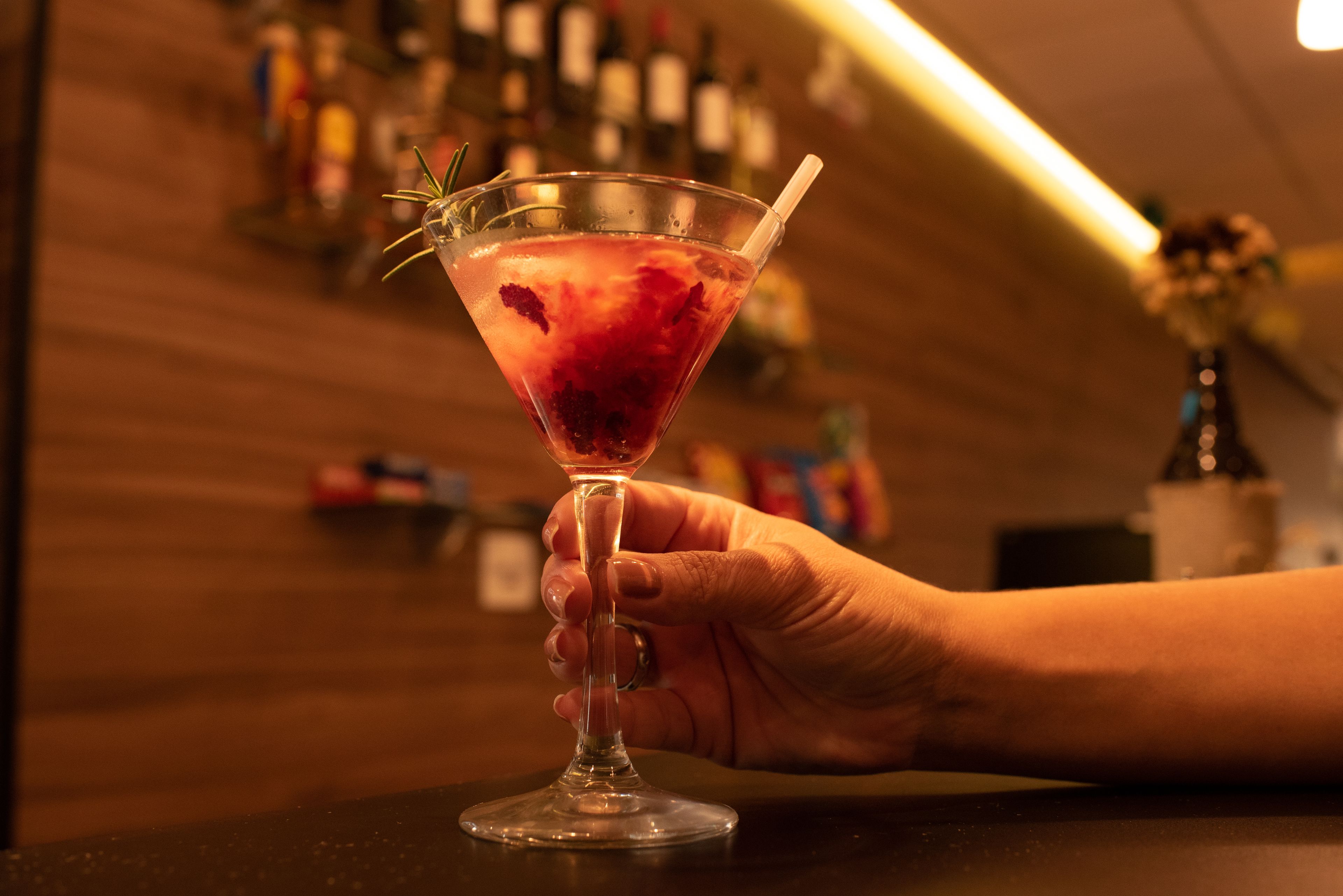 Une main tenant un verre à martini avec une paille et une garniture, le tout sur fond de bar avec des bouteilles en arrière-plan.