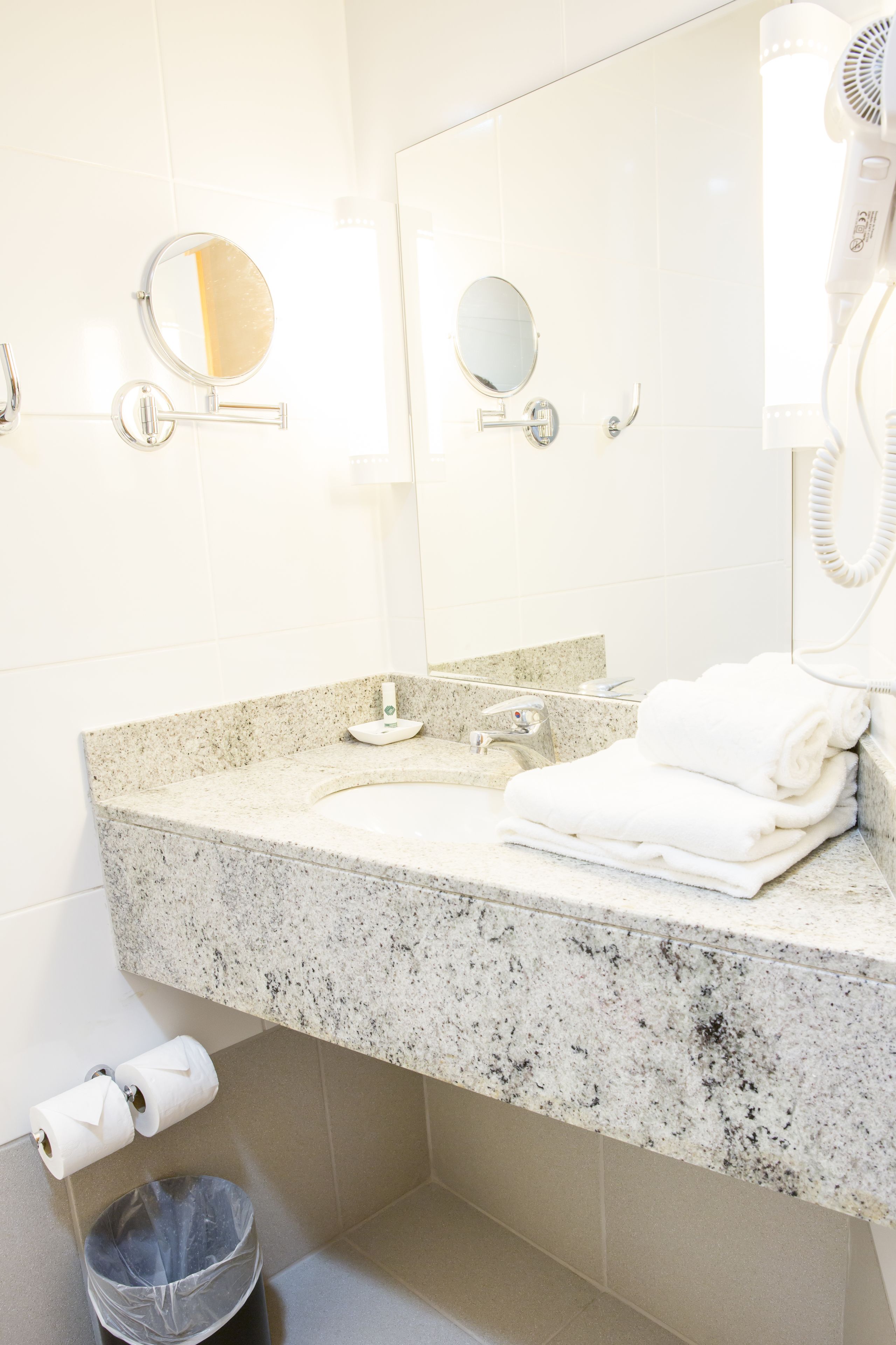 Une salle de bain avec un comptoir en granit, deux miroirs, un sèche-cheveux et une poubelle.