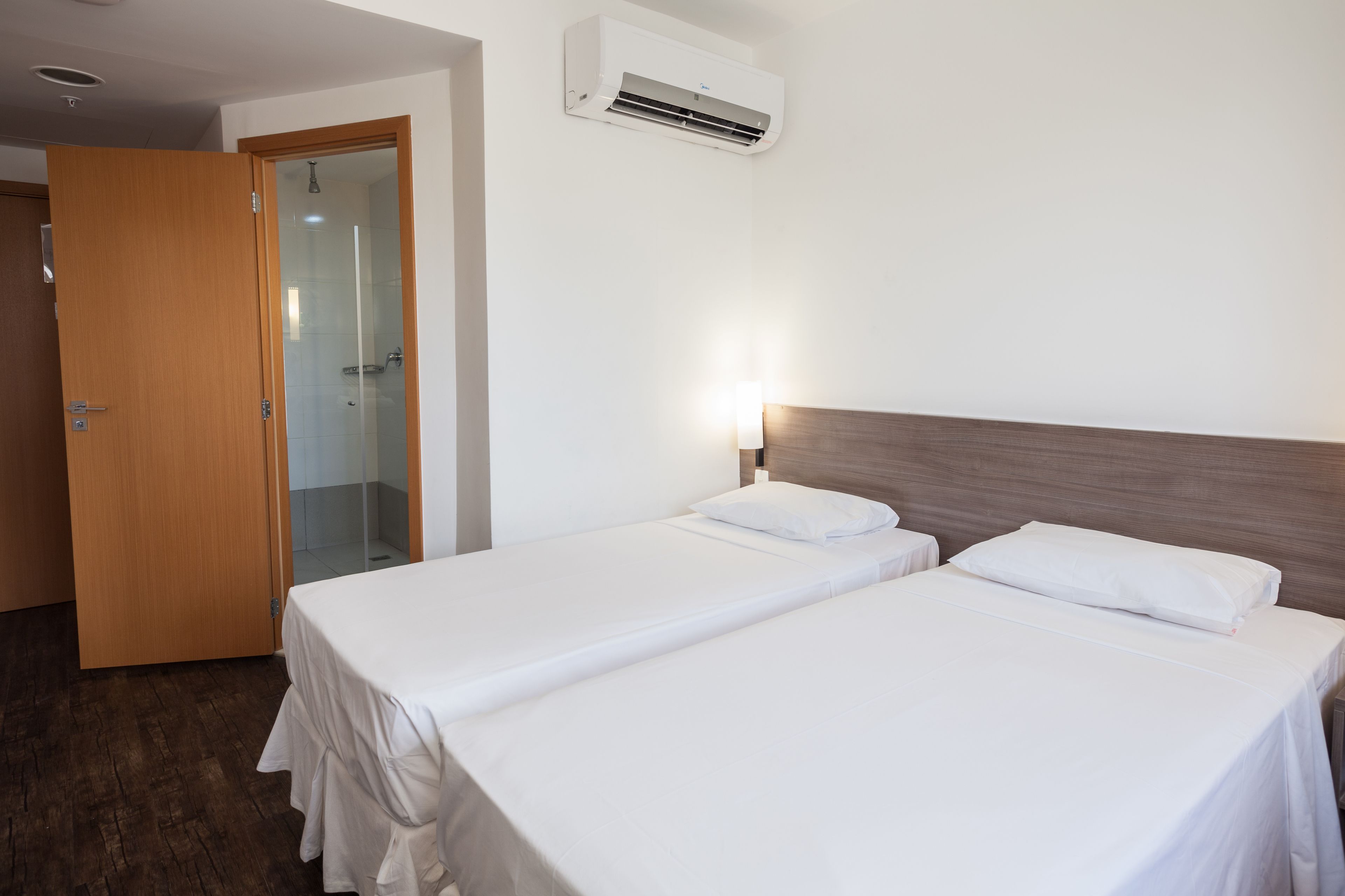 A hotel room with two beds, a wooden door, a glass shower enclosure, and an air conditioning unit.