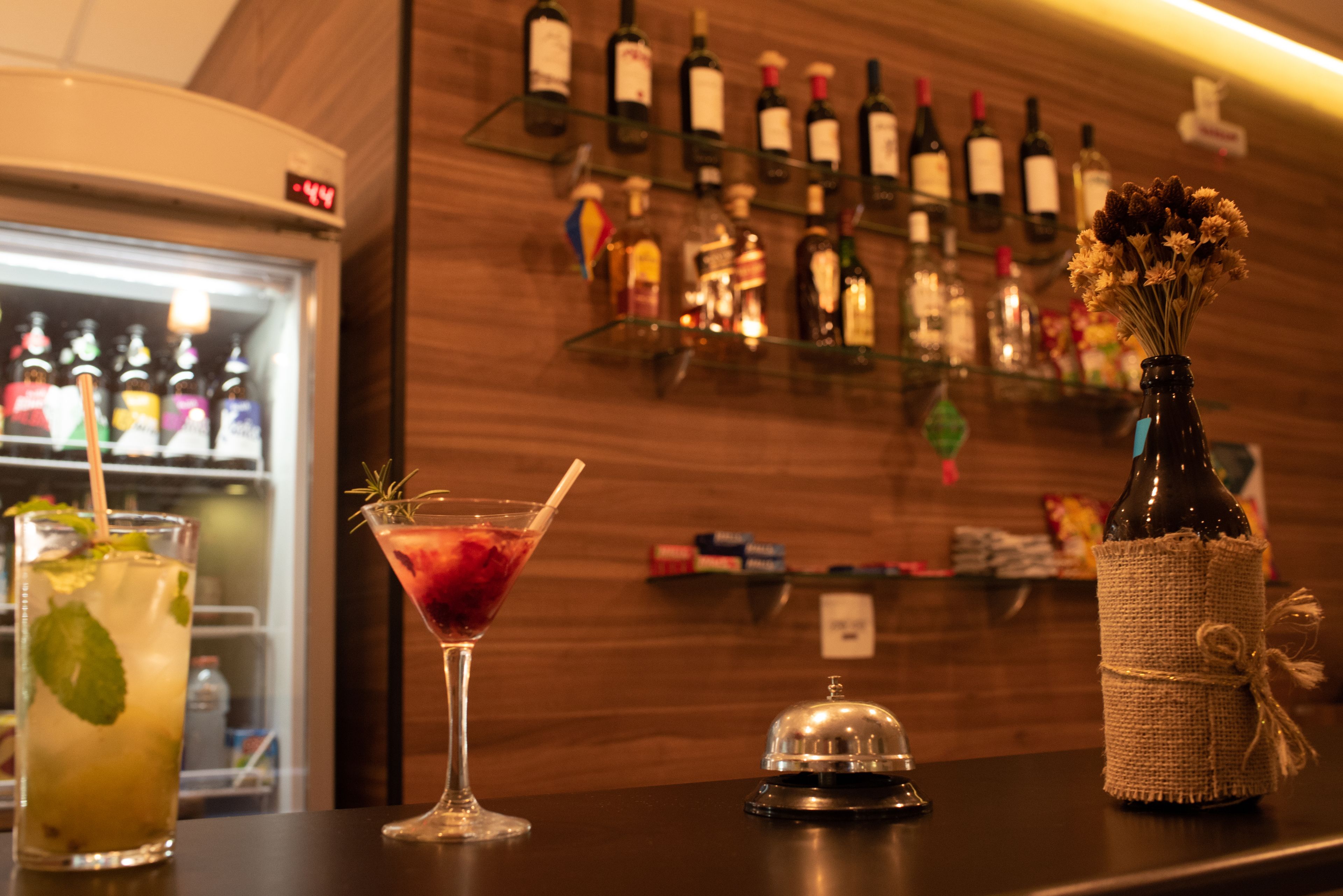 Un espace bar avec un assortiment de boissons en bouteille présentées sur des étagères, un réfrigérateur-vitrine et un vase de fleurs sur le comptoir.