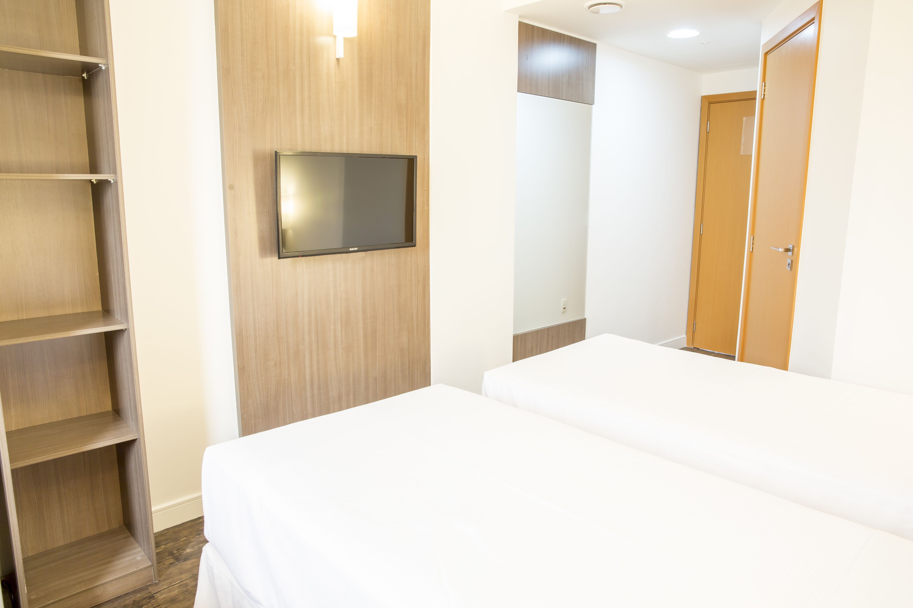 Una habitación de hotel con cama, televisor montado en la pared, armario de madera y una puerta.