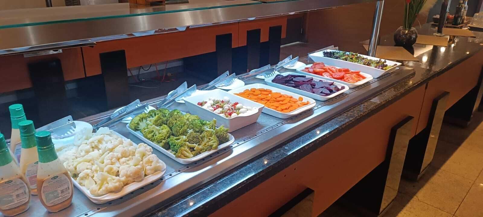 Buffet com diversos alimentos, incluindo brócolis, couve-flor, cenouras, tomates e outros vegetais.
