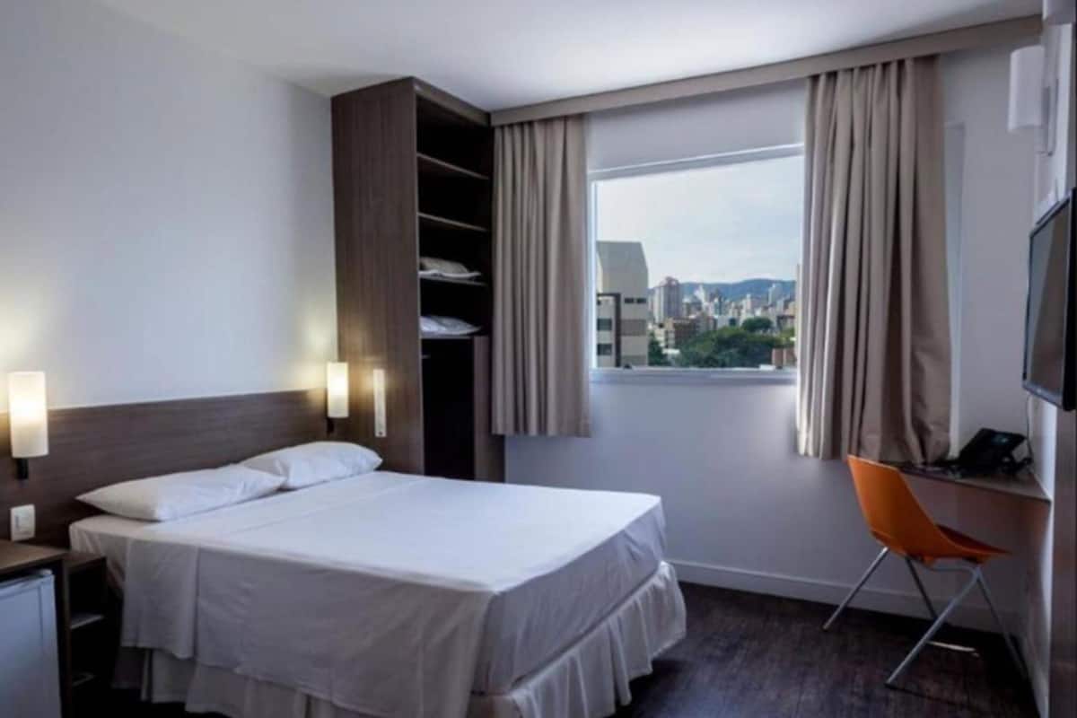 Une chambre d’hôtel avec un lit, un bureau, une chaise, une télévision et une fenêtre avec des rideaux.