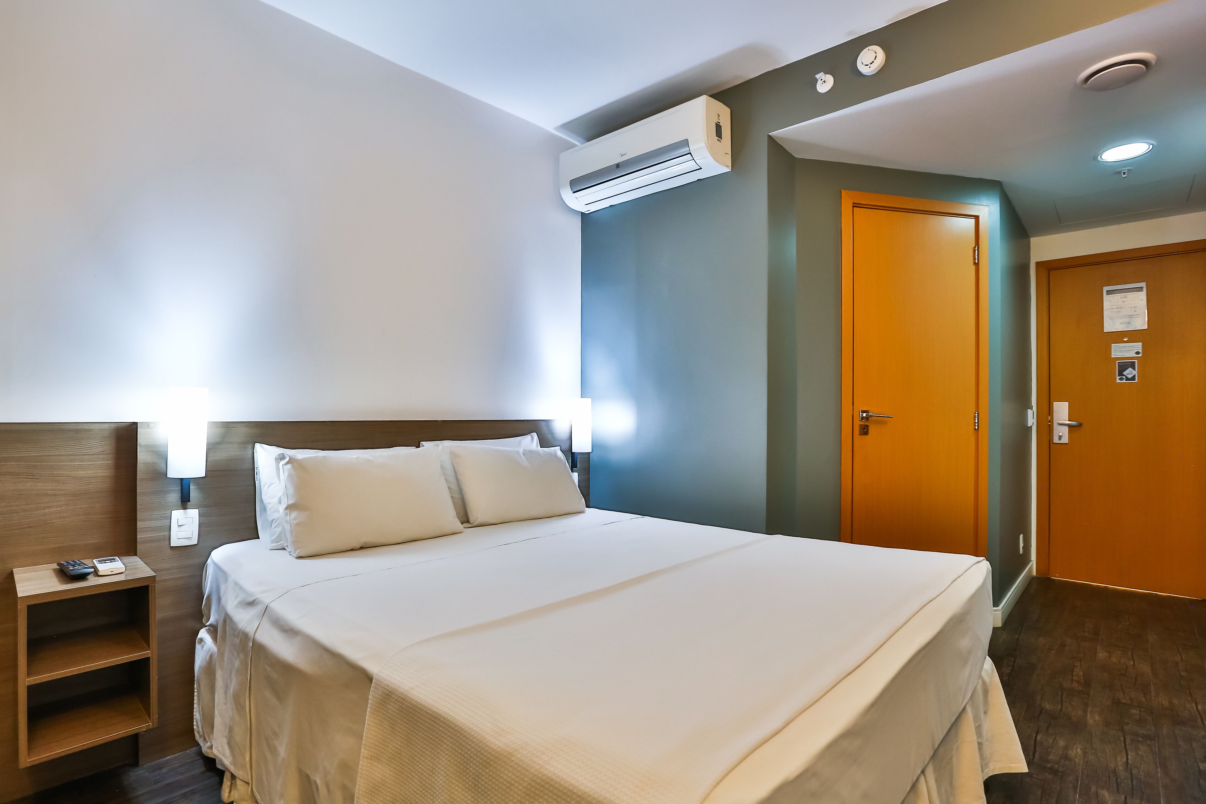 A hotel room with a bed, bedside table, nightstand, and an air conditioning unit.