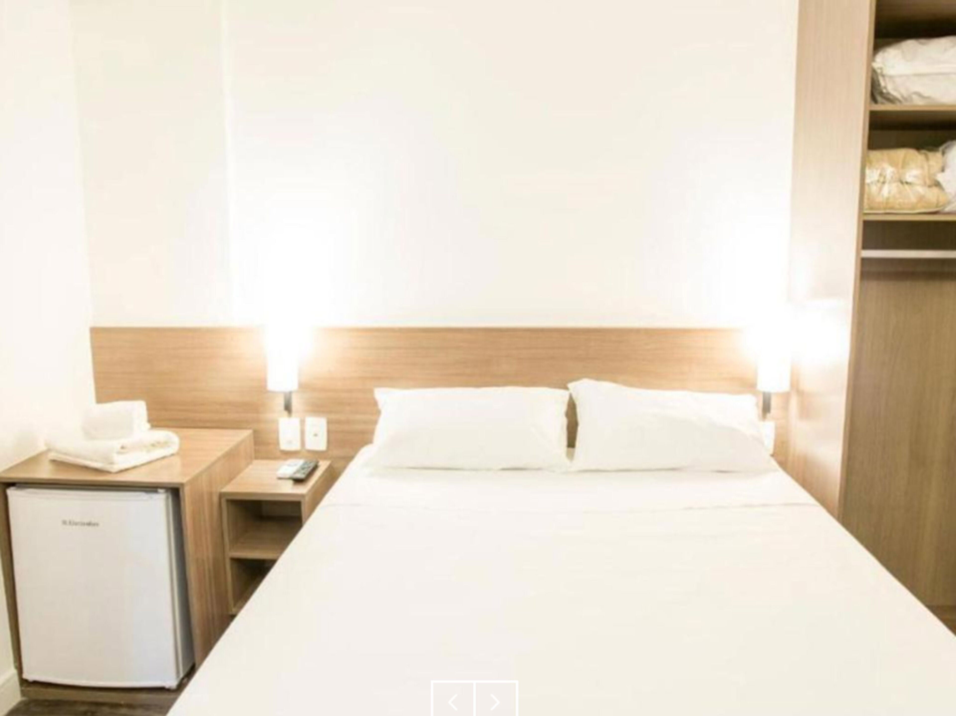 A hotel room with a bed, bedside tables, a minibar, and a wardrobe.