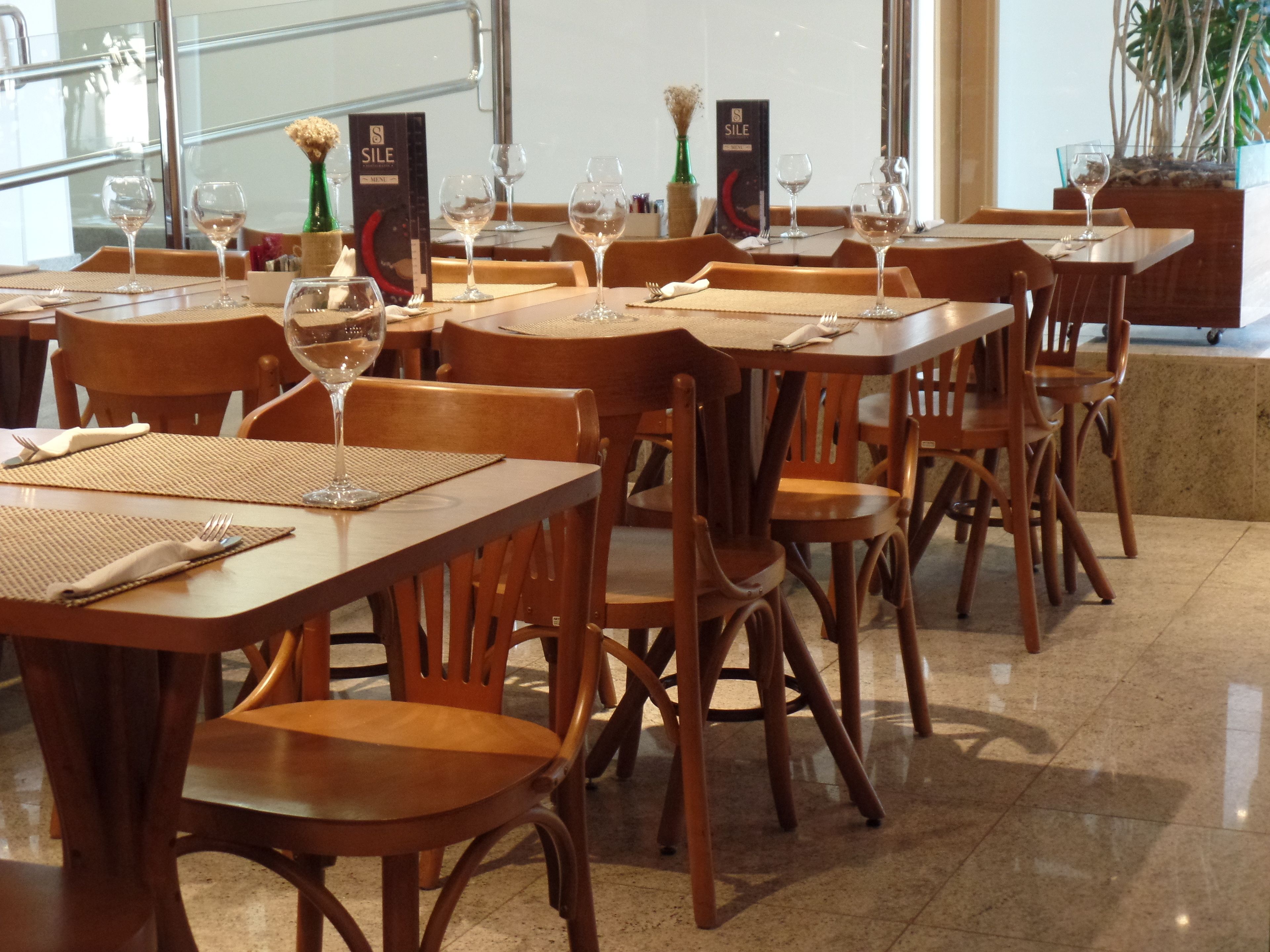 Um restaurante com mesas e cadeiras de madeira, copos de vinho, cardápios e um vaso com flores secas.