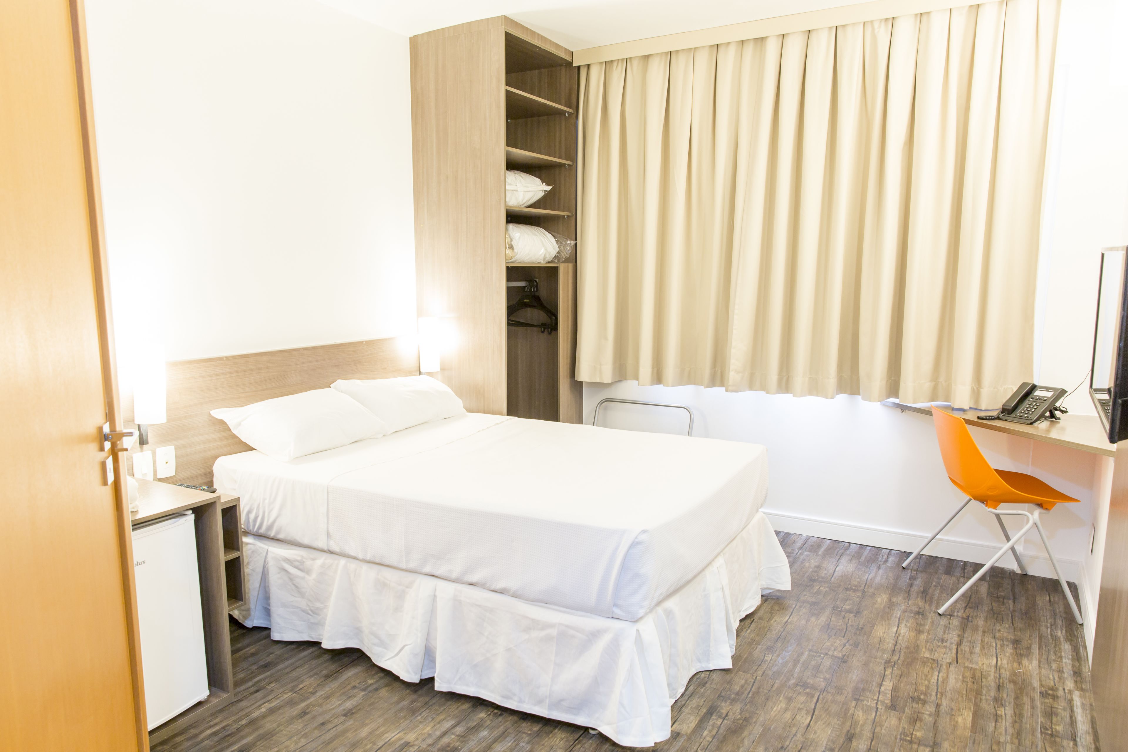 A hotel room with a bed, a desk with a chair, a wardrobe, and a window with curtains.