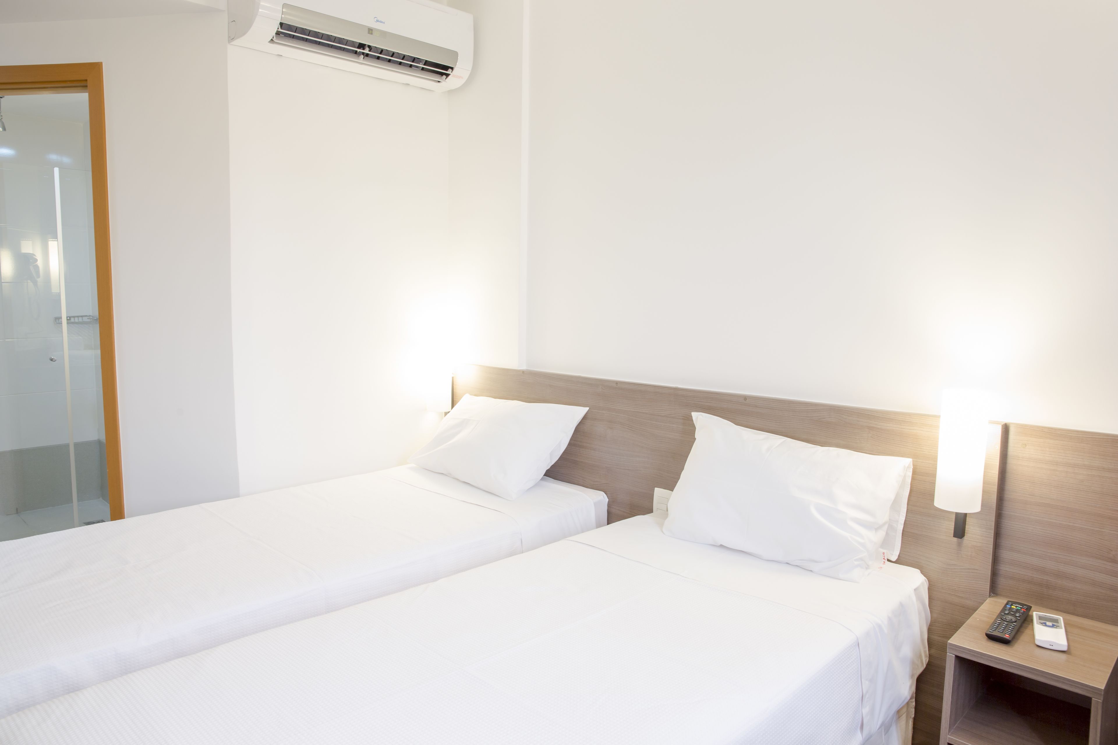 A hotel room with two single beds, a bedside table with a remote control, and an air conditioning unit on the wall.
