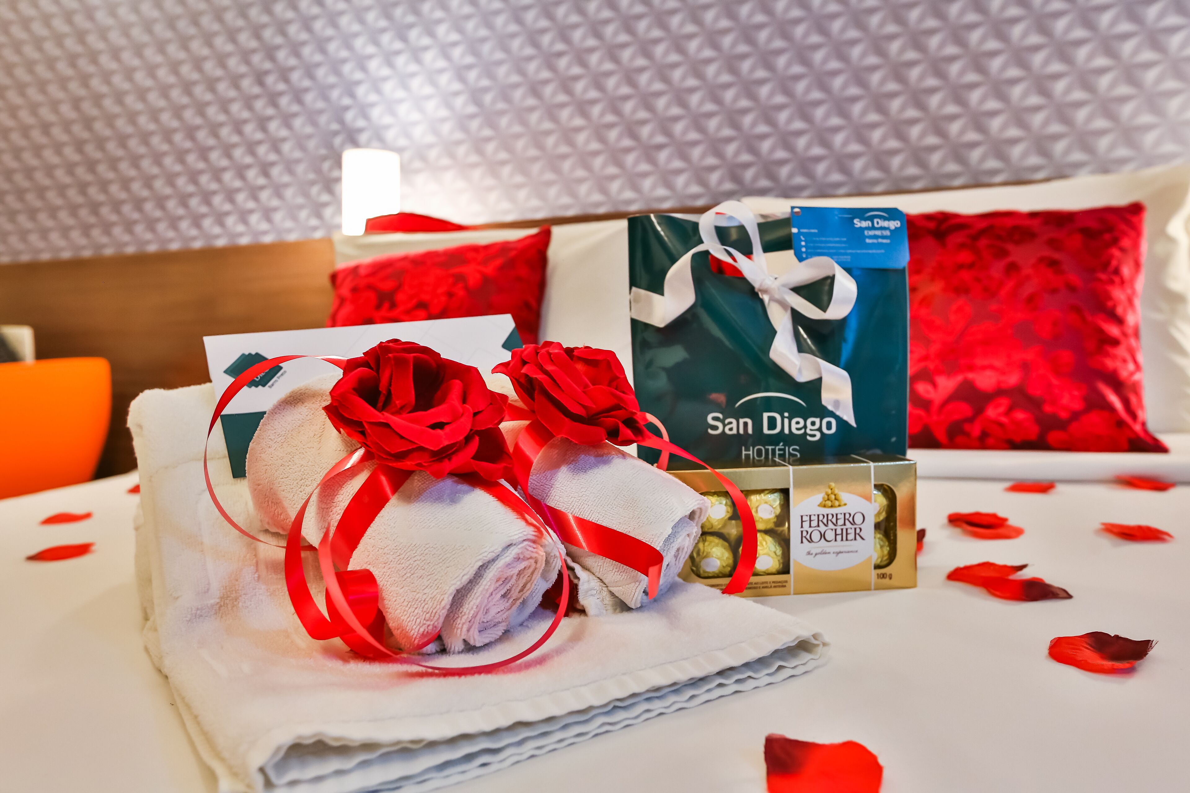 Una cama cuidadosamente tendida con una toalla blanca enrollada, adornos de rosas rojas y una caja de regalo con la etiqueta 'San Diego Hotels'.