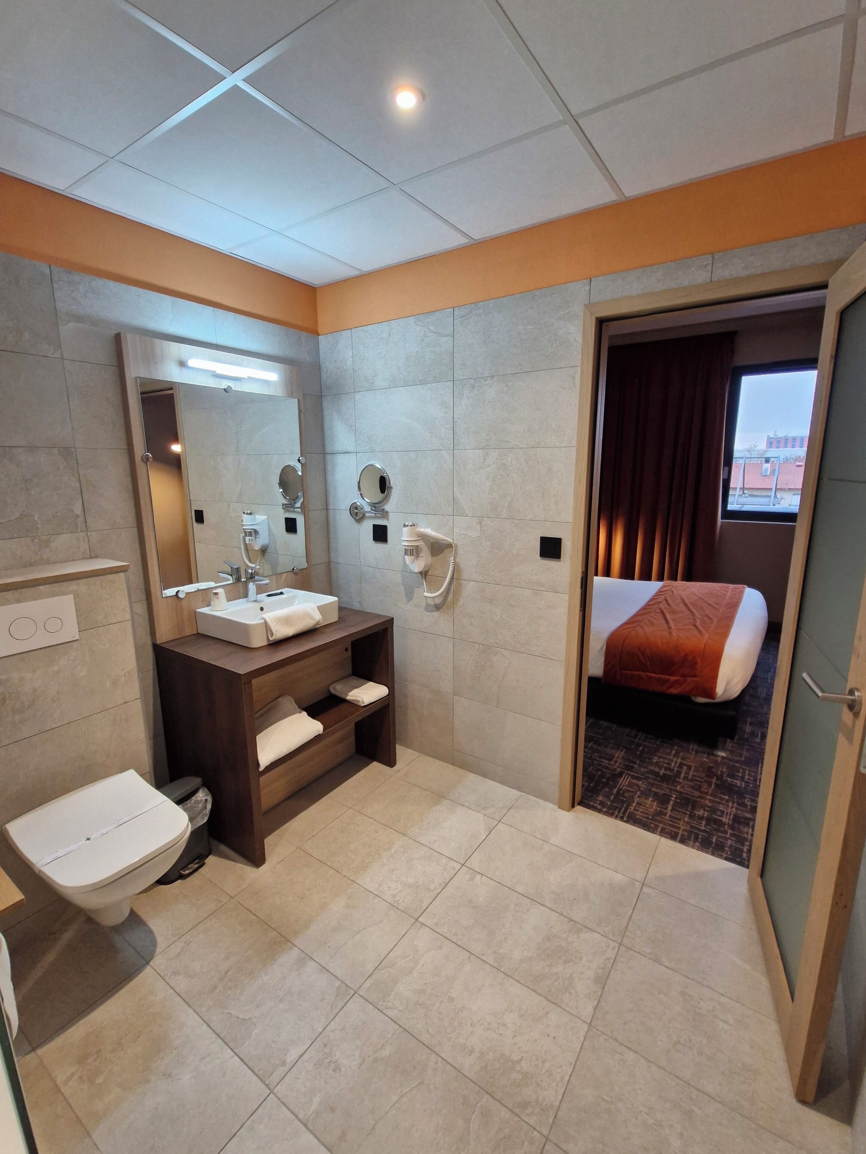 A hotel bathroom with a toilet, sink, mirror, and a hairdryer on the wall.
