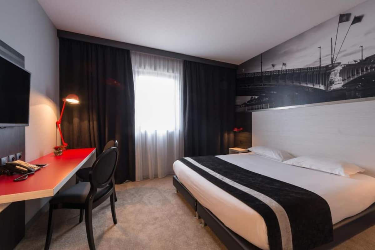 Una habitación de hotel con cama, escritorio, silla, lámpara y ventana con cortinas.