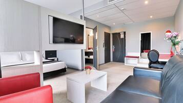 A modern hotel room with a bed, a TV, a desk, and seating areas.