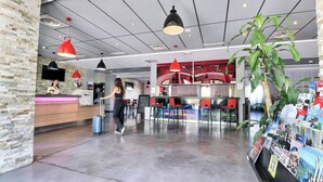 Et hotel lobby med en receptionsdisk, røde hængelamper, en stor stueplante og en brochurestander.