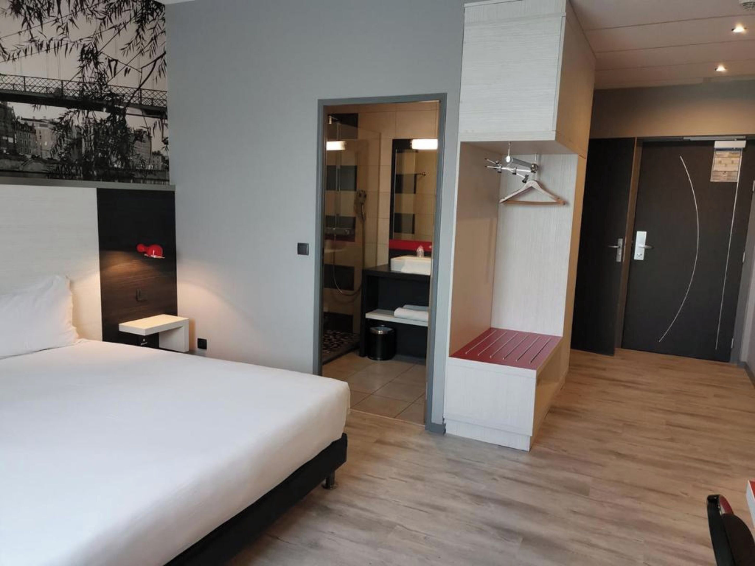 Une chambre d’hôtel moderne avec un grand lit, une table de chevet, une lampe suspendue, une armoire et une salle de bain visible à travers une porte ouverte.