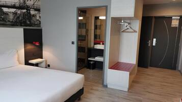 Une chambre d’hôtel moderne avec un grand lit, une table de chevet, une lampe suspendue, une armoire et une salle de bain visible à travers une porte ouverte.