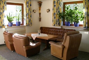 Un salon avec un canapé en cuir marron, deux fauteuils, une table basse en bois et deux plantes en pot posées sur les rebords des fenêtres.