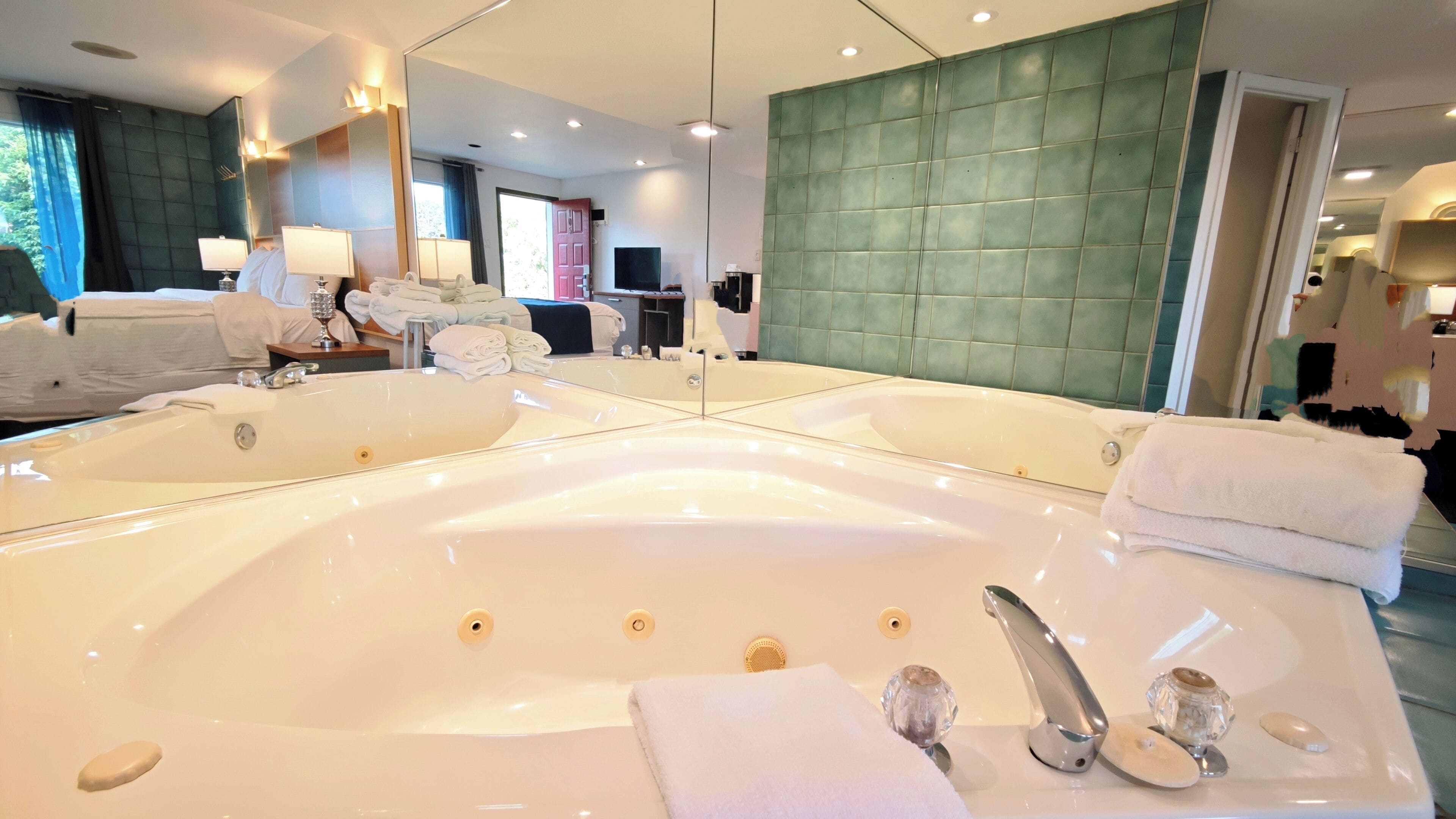A large, white bathtub with a mirror above it, a green upholstered wall, and a door leading to another room.