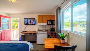 A compact kitchen with a microwave, stove, and sink, a dining table with a chair, a TV mounted on the wall, and a window with a view of the outside.