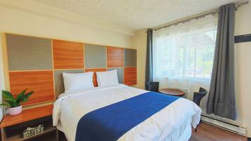 A hotel room with a large bed, a wooden headboard, a small table, a chair, and a potted plant.