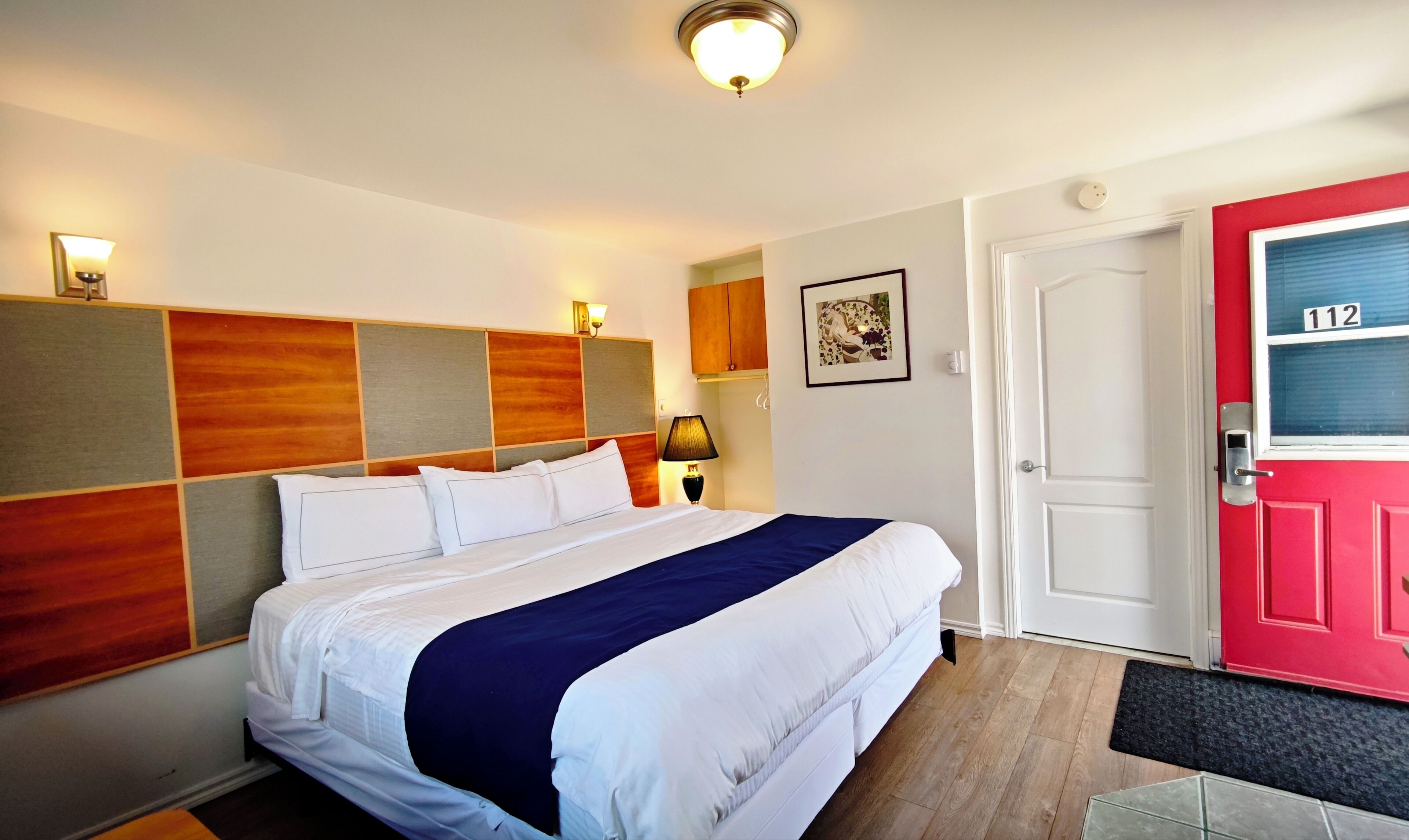 Habitación de hotel con cama, mesita de noche, lámpara, puerta con un número y aplique de pared.