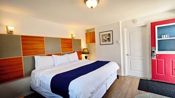 Habitación de hotel con cama, mesita de noche, lámpara, puerta con un número y aplique de pared.