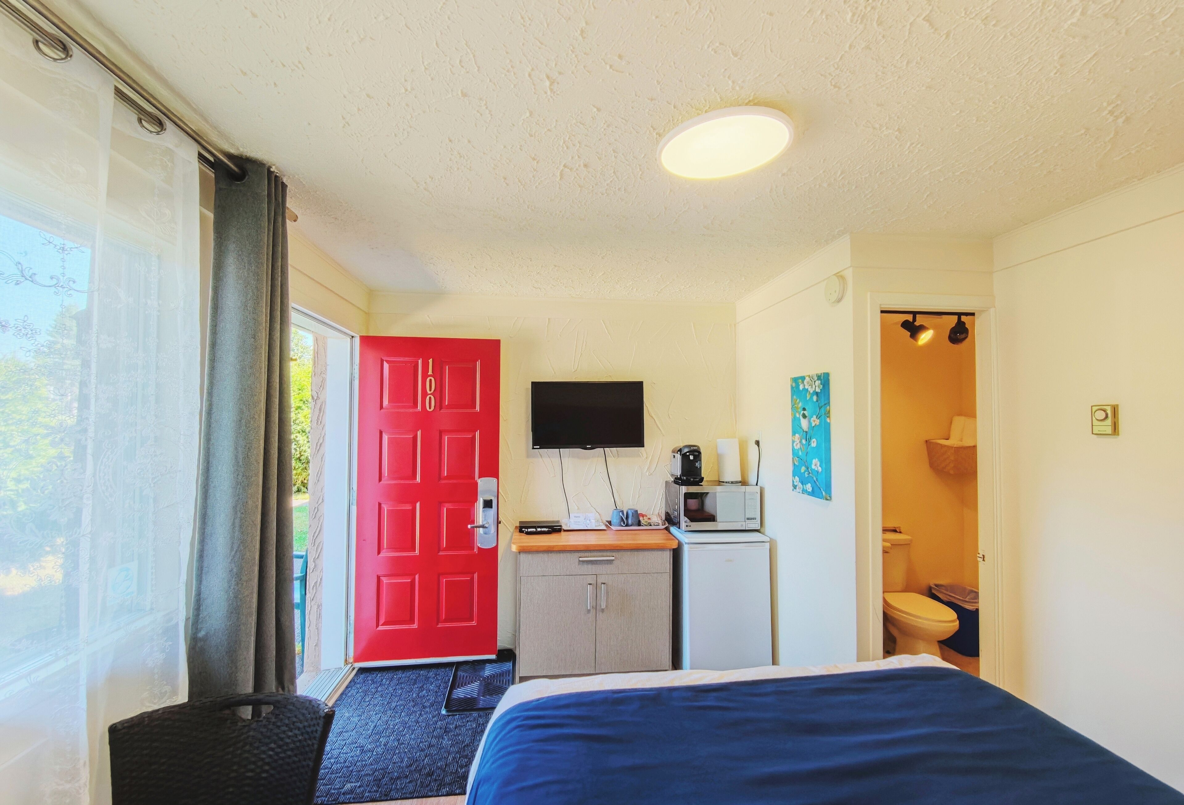 Quarto com cama, escrivaninha com televisão, pequeno espaço de cozinha e banheiro.