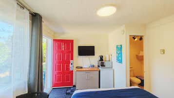 Quarto com cama, escrivaninha com televisão, pequeno espaço de cozinha e banheiro.