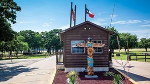 A small wooden cabin with a totem pole in front, surrounded by a landscaped garden with small plants and a paved road in the background. - Lake Tawakoni RV Campground (Point)