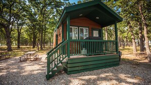 A small cabin with a green porch and a wooden deck, surrounded by trees in a park setting. - Lake Tawakoni RV Campground (Point)