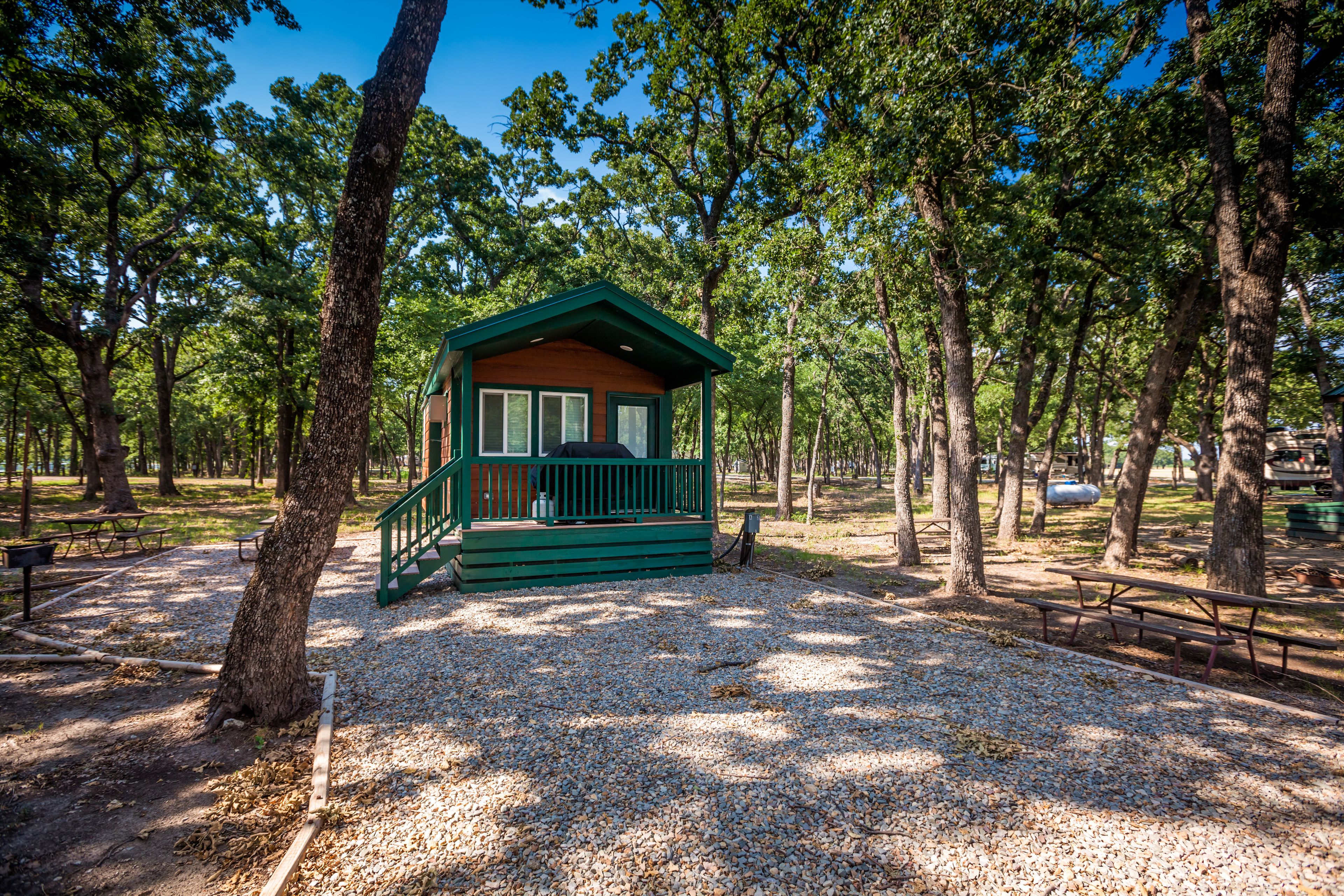 Een klein houten huisje met een groen dak en een houten terras, omgeven door bomen in een park.