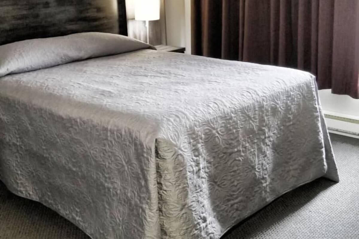 A neatly made bed with a grey bedspread, a bedside lamp, and a window with curtains.