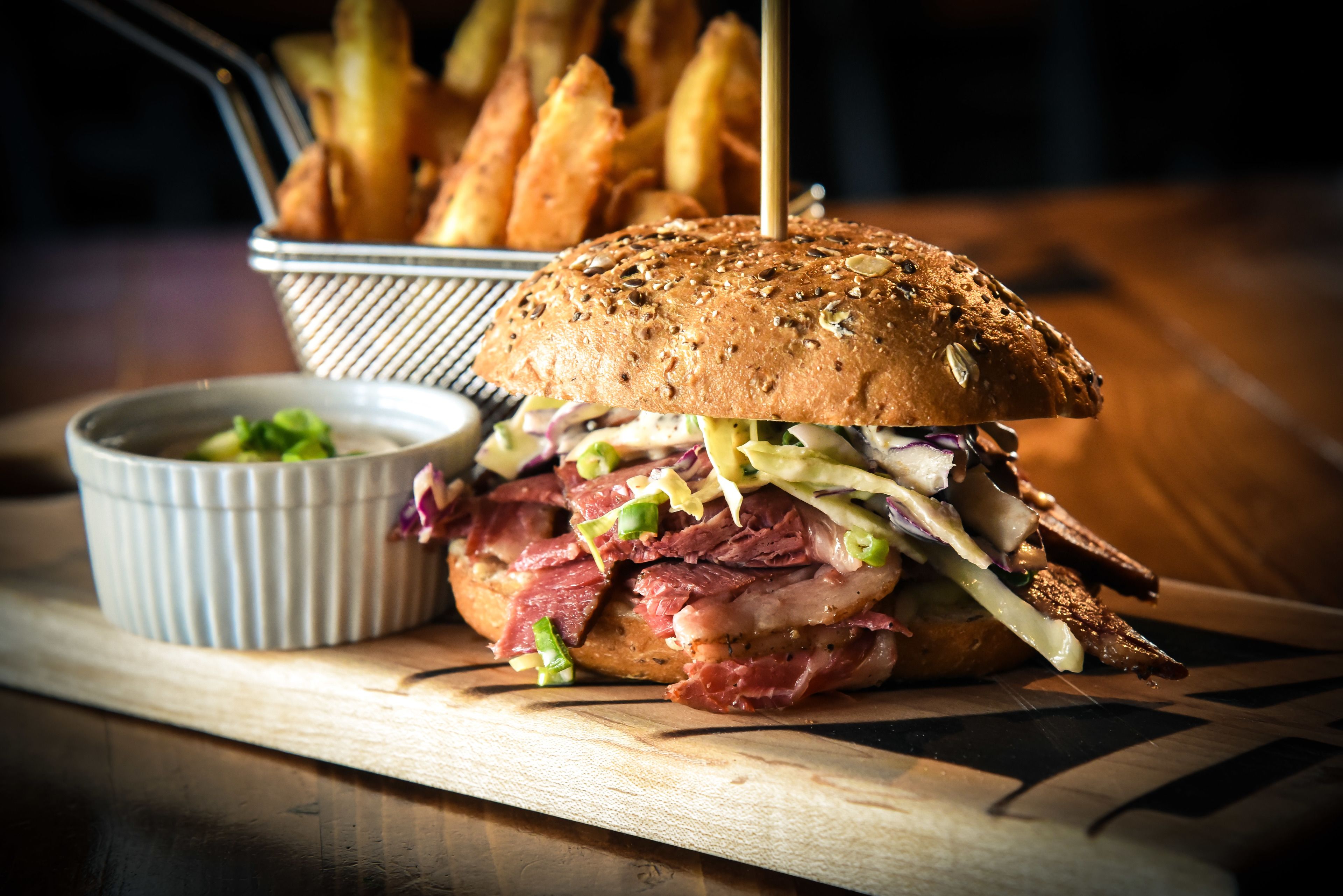 A sandwich with a bun, meat, and vegetables, accompanied by a small bowl of sauce and a basket of fries.