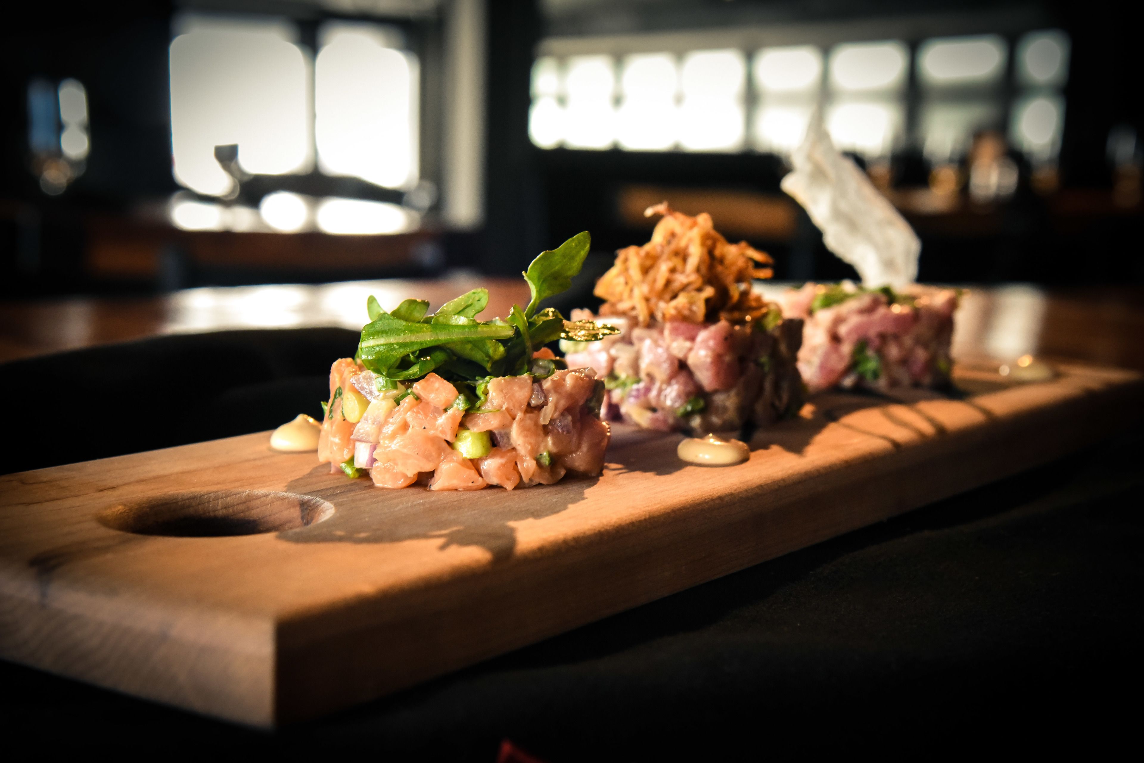 A wooden board with three sushi pieces, garnished with green leaves and sesame seeds, in a dimly lit setting.