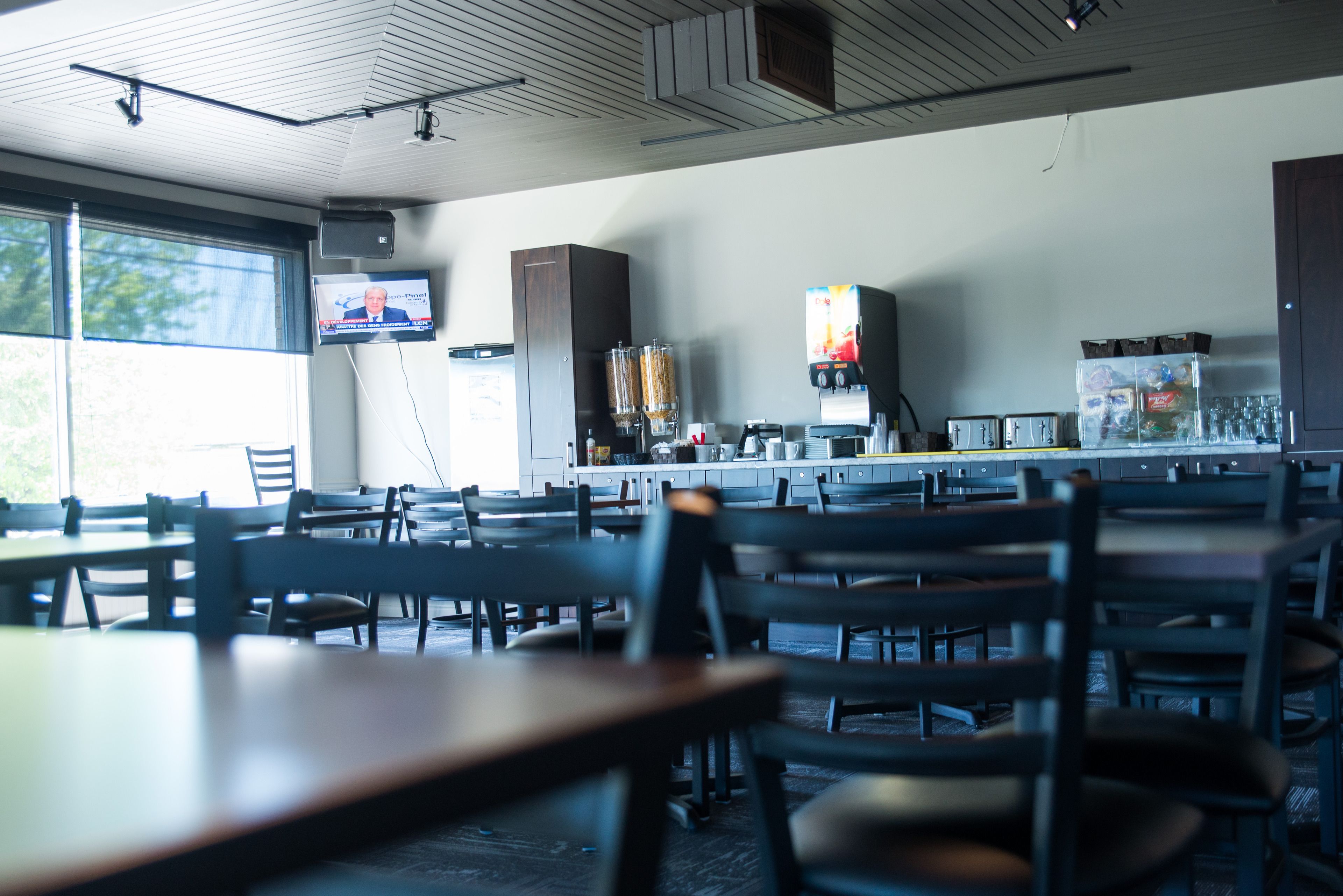A restaurant with multiple tables and chairs, a television mounted on the wall, and a kitchen area visible in the background.