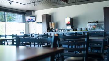 A restaurant with multiple tables and chairs, a television mounted on the wall, and a kitchen area visible in the background.