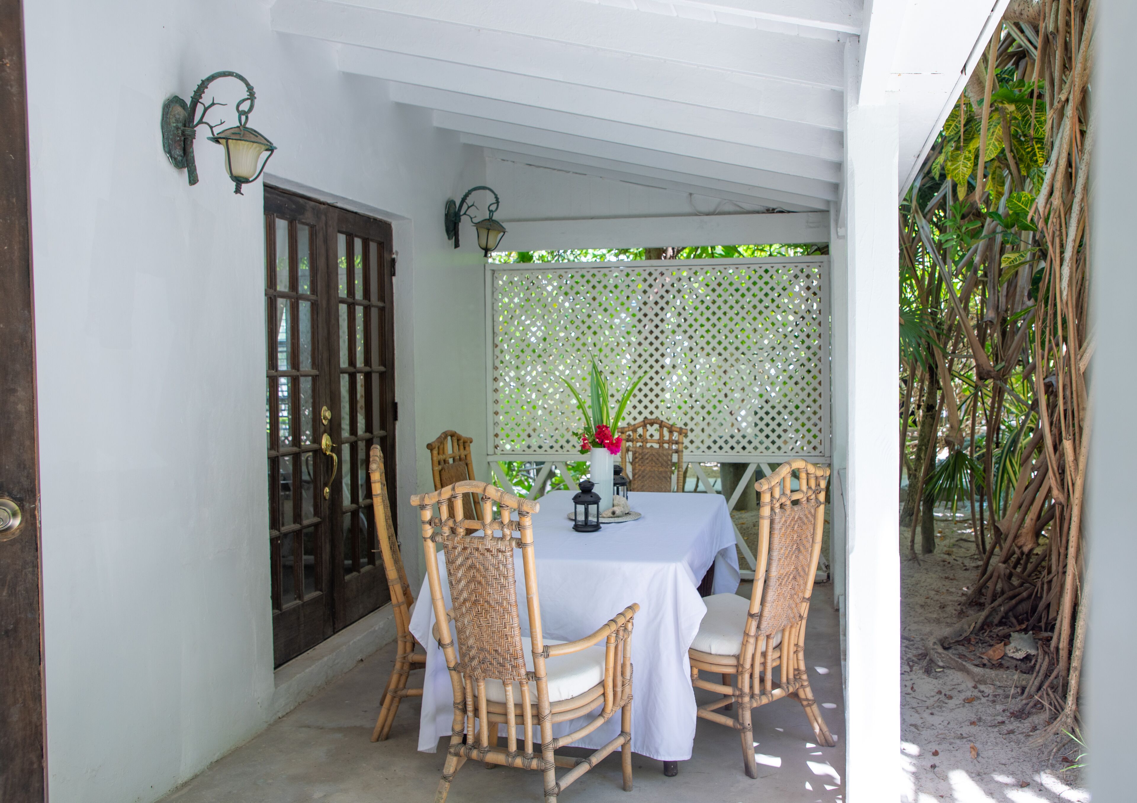 A covered outdoor dining area with a table set for four, surrounded by wicker chairs, a white tablecloth, and a decorative vase with flowers.