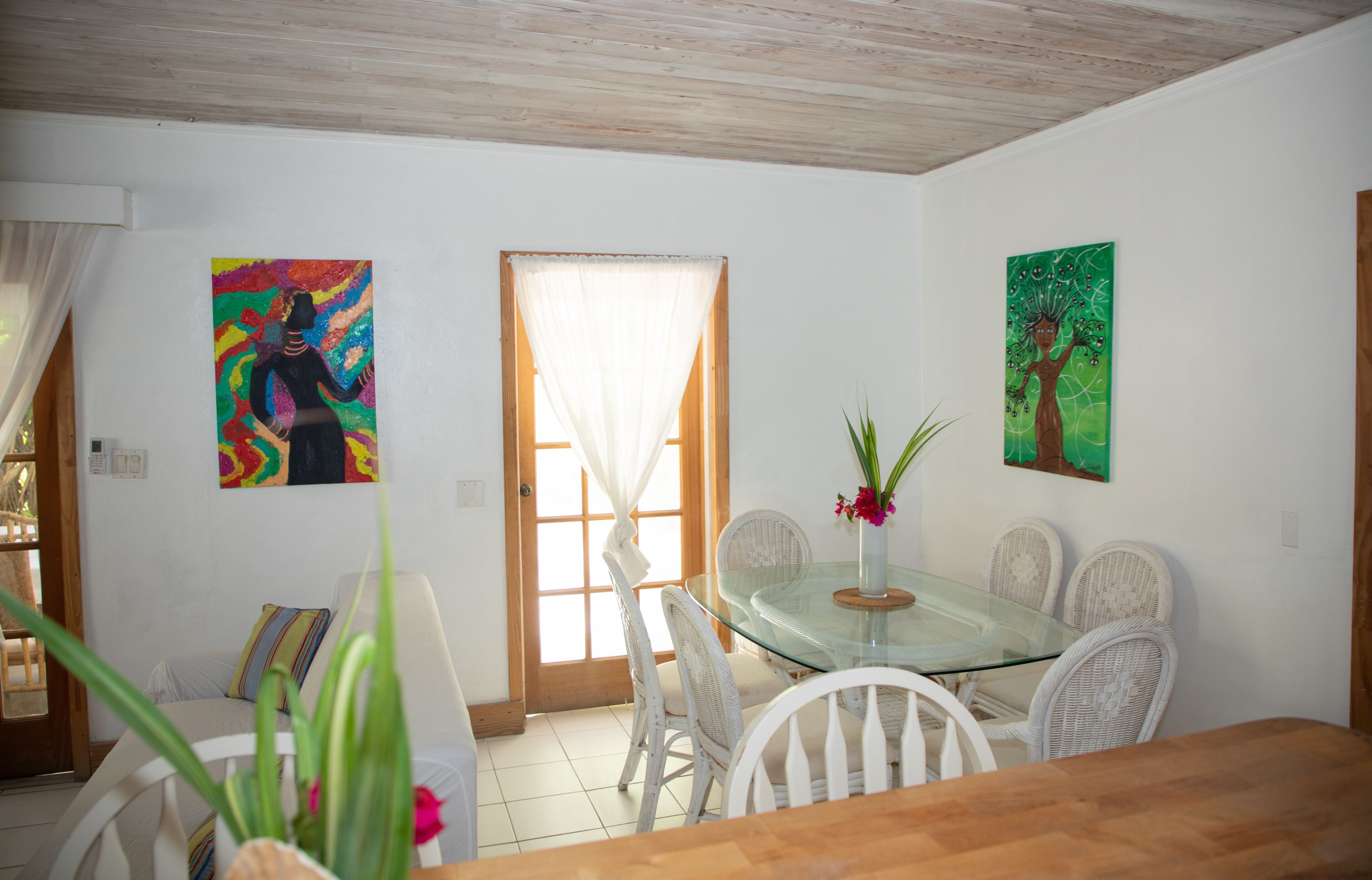 Un comedor con una mesa de vidrio, sillas de mimbre blanco y un florero con flores. Hay dos cuadros en las paredes, uno con un árbol y otro con un diseño abstracto y colorido.