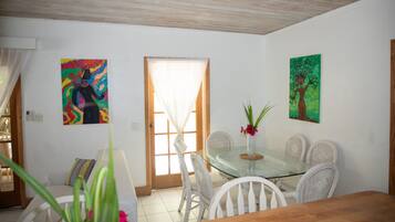 Un comedor con una mesa de vidrio, sillas de mimbre blanco y un florero con flores. Hay dos cuadros en las paredes, uno con un árbol y otro con un diseño abstracto y colorido.
