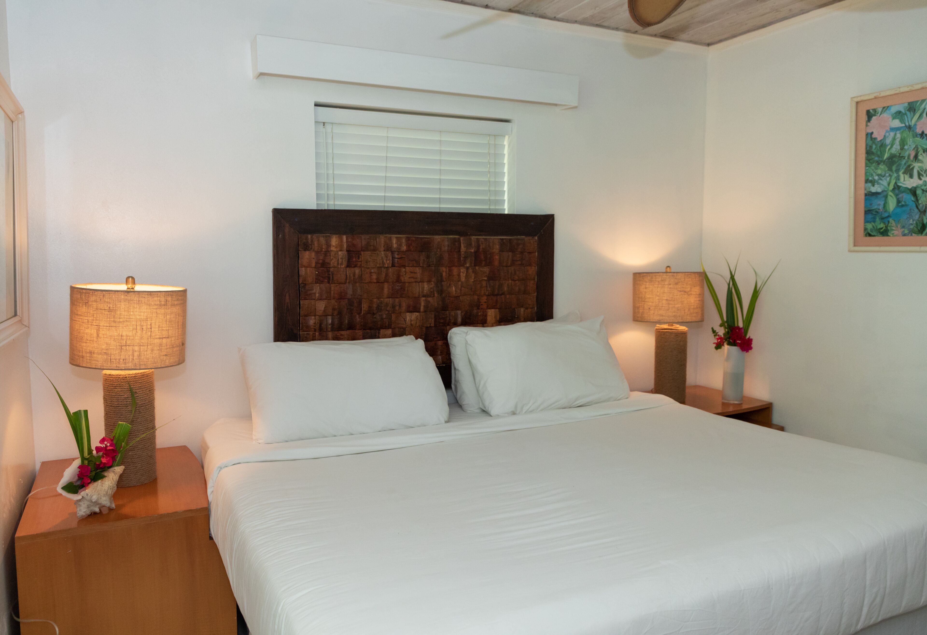 A neatly made bed with white linens, two bedside lamps, a wooden headboard, and a window with blinds.