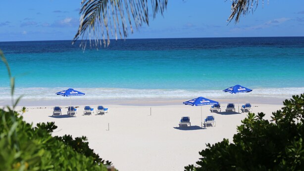 A beach with white sand, clear blue water, and several blue umbrellas. - Tingum Village Hotel (Dunmore Town)